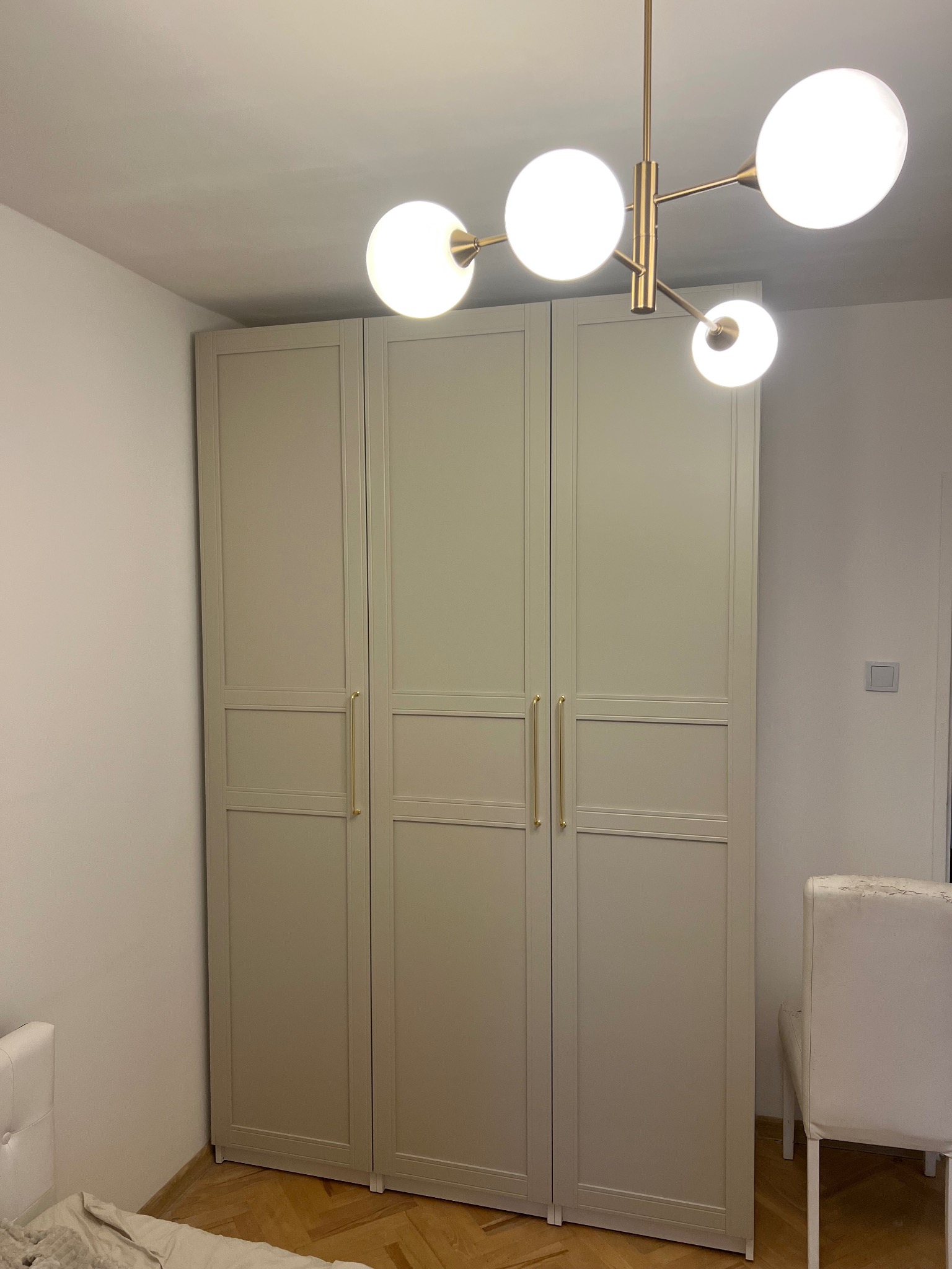 Trzydrzwiowa szafa w odcieniach beżu z złotymi uchwytami, oświetlona lampą sufitową z kulistymi kloszami, w minimalistycznym wnętrzu.