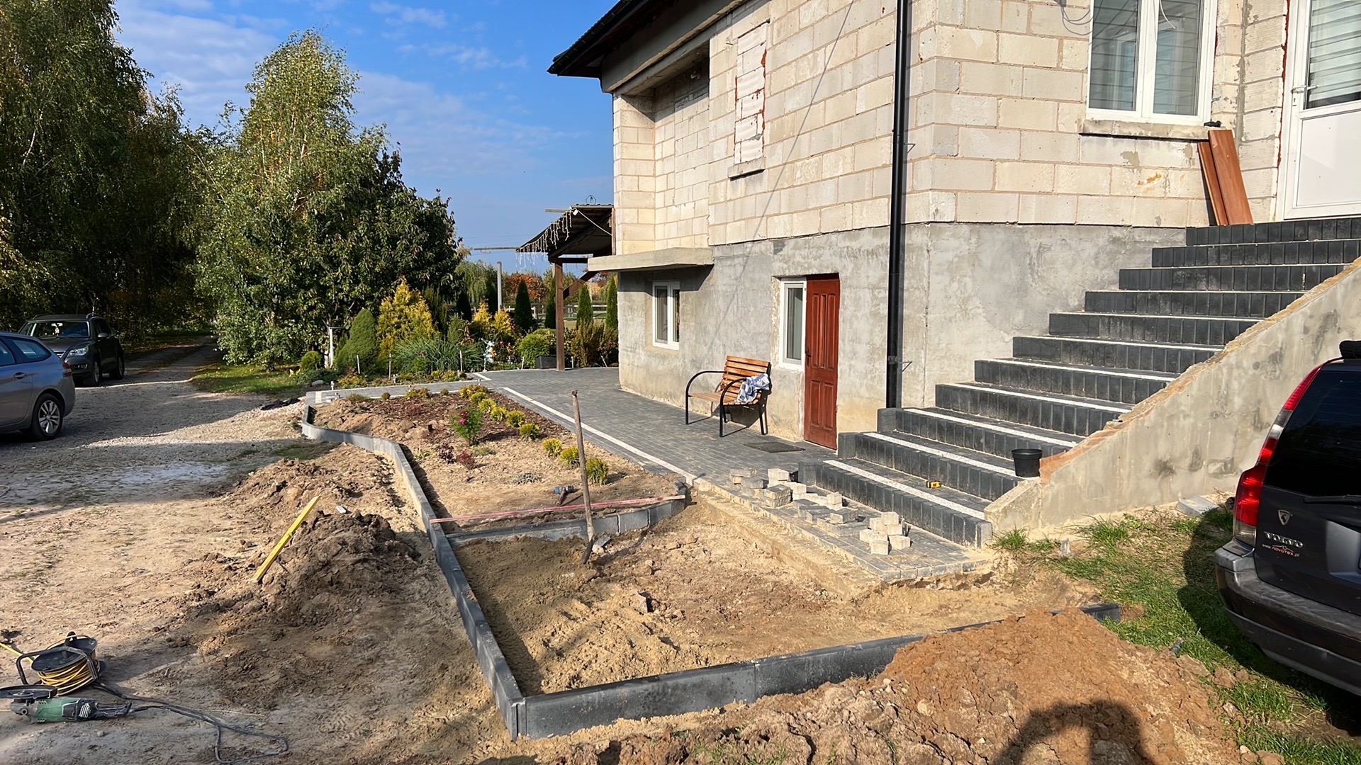 Prace ziemne przed domem: przygotowanie gruntu pod rabaty kwiatowe z obrzeżami, widoczne schody wejściowe i fragment elewacji budynku. Narzędzia ogrodnicze na pierwszym planie.