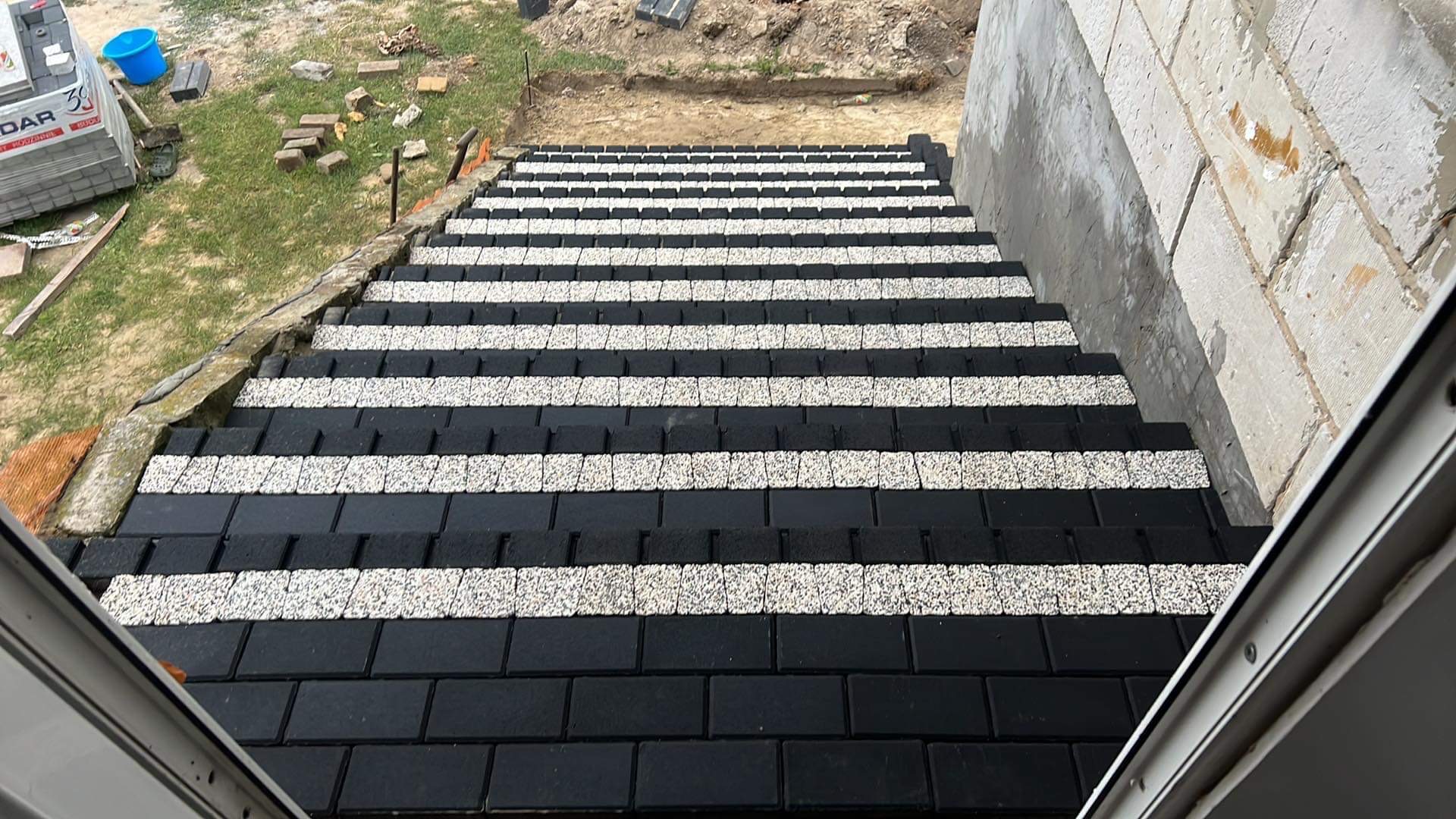 Schody zewnętrzne wyłożone czarno-białą kostką brukową, tworzącą wzór pasów. Widok z góry, ukazujący detale ułożenia i kontrast kolorów.