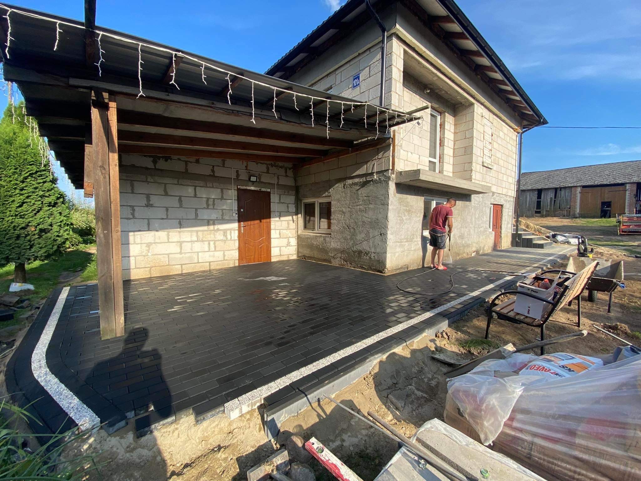 Nowa, czarna kostka brukowa ułożona na tarasie domu z oświetleniem girlandą. Mężczyzna w czerwonej koszulce przy ścianie. Widoczne narzędzia i worki z materiałami.