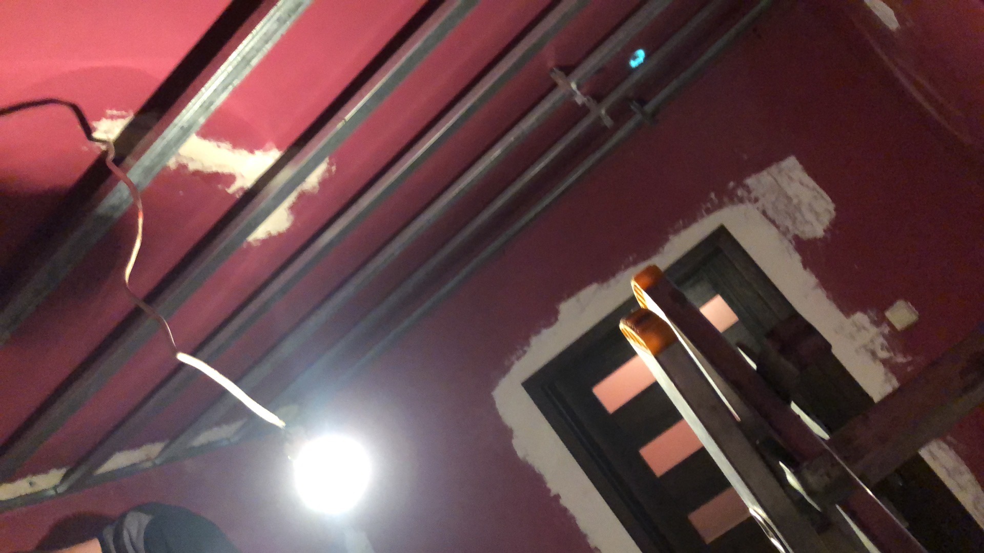 Czerwona ściana w trakcie remontu z widocznym stelażem sufitowym, fragmentem drabiny i niedomalowanym obramowaniem drzwi, oświetlona lampą.