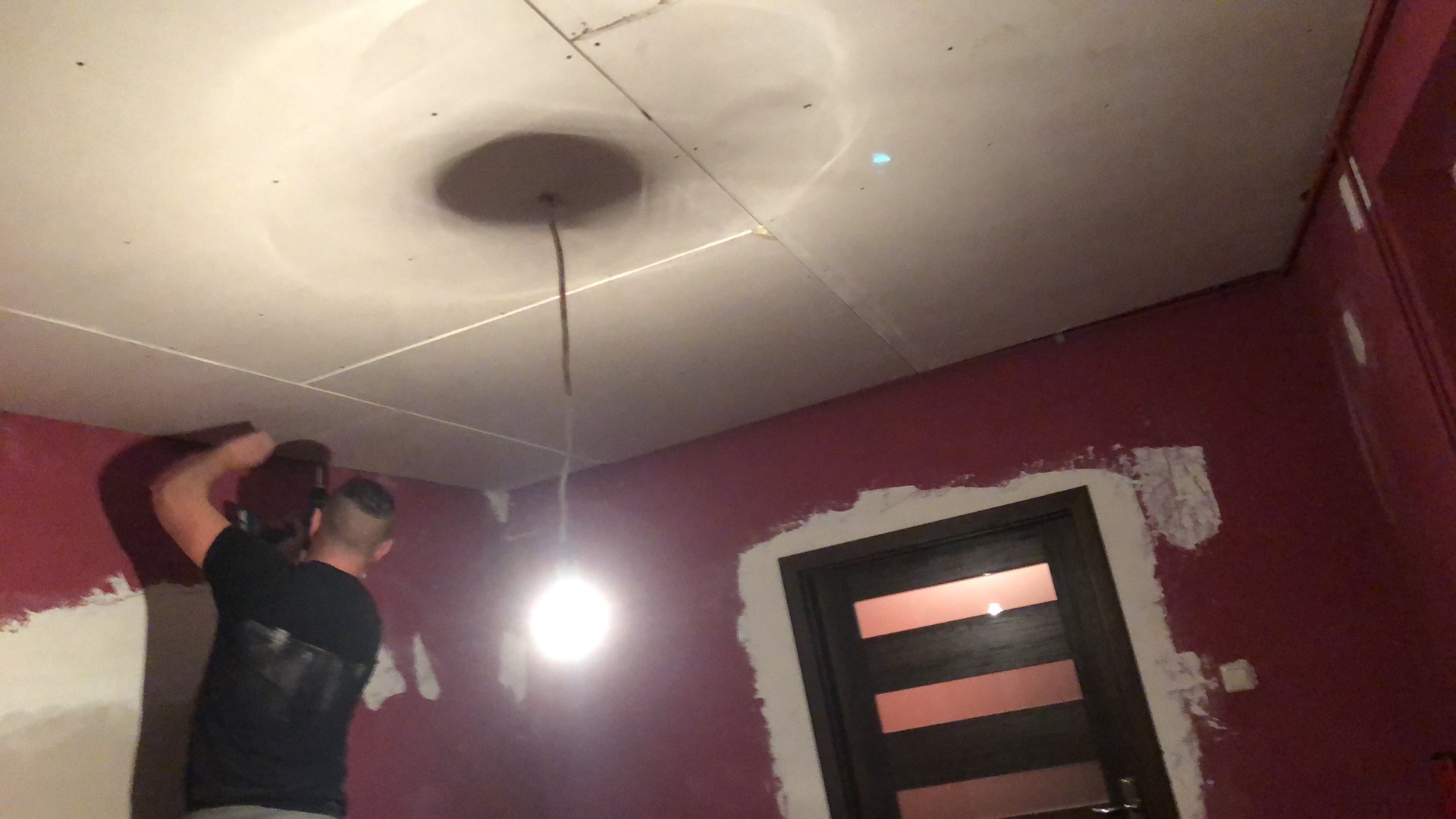 Montaż płyt karton-gips na suficie w pokoju z bordowymi ścianami. Widoczny pracownik podczas pracy i fragment drzwi. Sufit z okrągłym elementem dekoracyjnym.