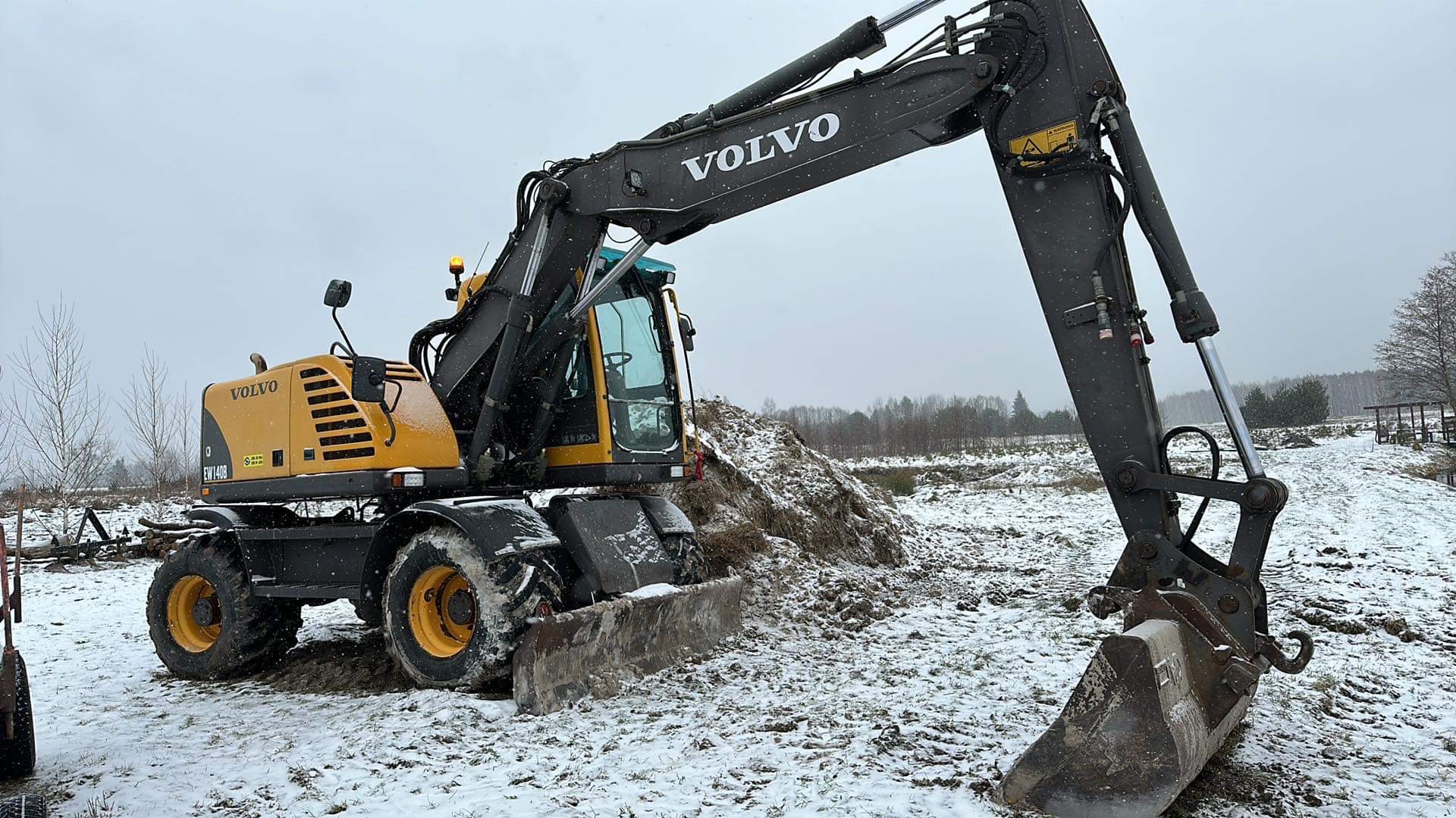 Żółto-czarna koparka Volvo na śnieżnym terenie z usypaną ziemią w tle, pochmurny dzień. Widoczne detale konstrukcji i łyżka koparki.