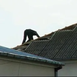 Pracownik w ciemnym kombinezonie na dachu z eternitu, demontaż pokrycia dachowego, widoczne deski i elementy konstrukcji dachu. Ujęcie z dołu, pochmurne niebo.