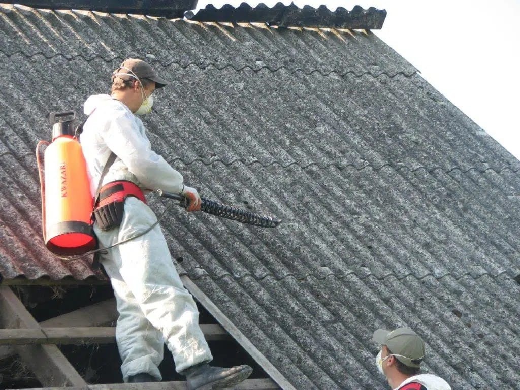 Pracownik w kombinezonie ochronnym, z opryskiwaczem na plecach, zabezpiecza eternitowy dach. Ujęcie z dołu, widoczna struktura dachu i drugi pracownik w tle.