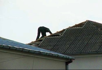 Pracownik w ciemnym kombinezonie na dachu z eternitu, demontaż pokrycia dachowego, widoczne deski i elementy konstrukcji dachu. Ujęcie z dołu, pochmurne niebo.