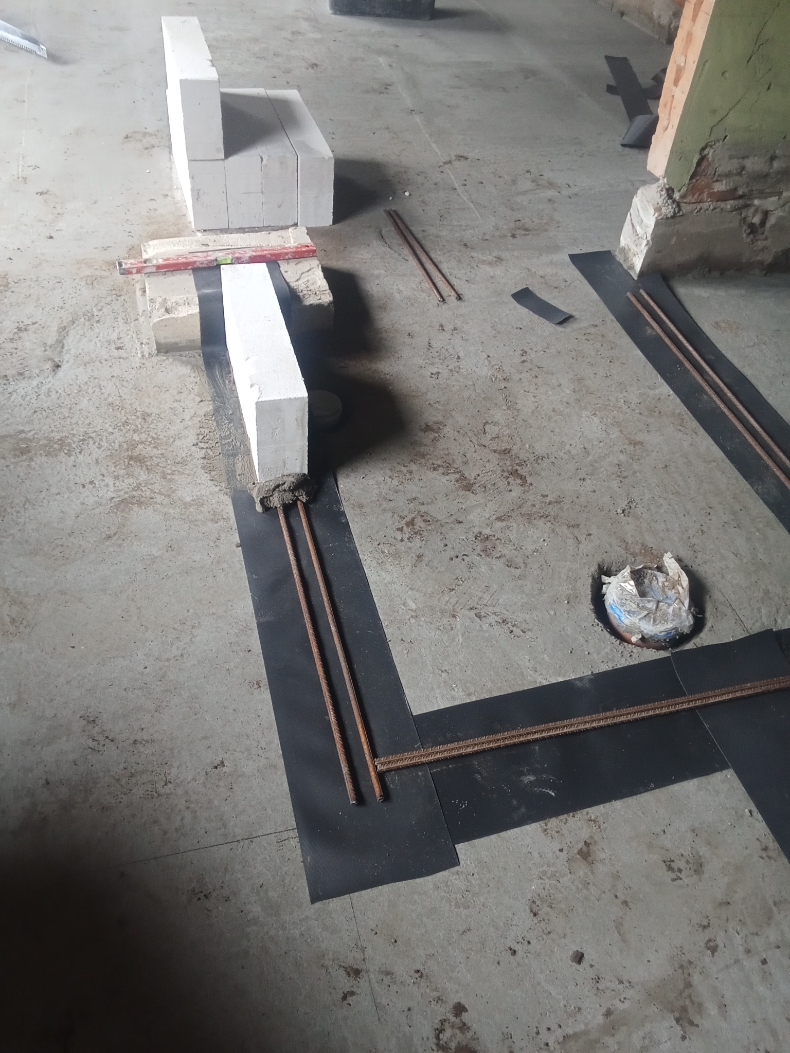 Wznoszenie ściany z bloczków betonowych na fundamencie z hydroizolacją i zbrojeniem. Widoczny poziomica i fragment starej ściany.