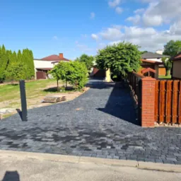 Ułożona, ciemnoszara kostka brukowa prowadząca wzdłuż ogrodzenia z cegły i drewna. Widoczne drzewa, domy w tle i fragment nieba z chmurami.