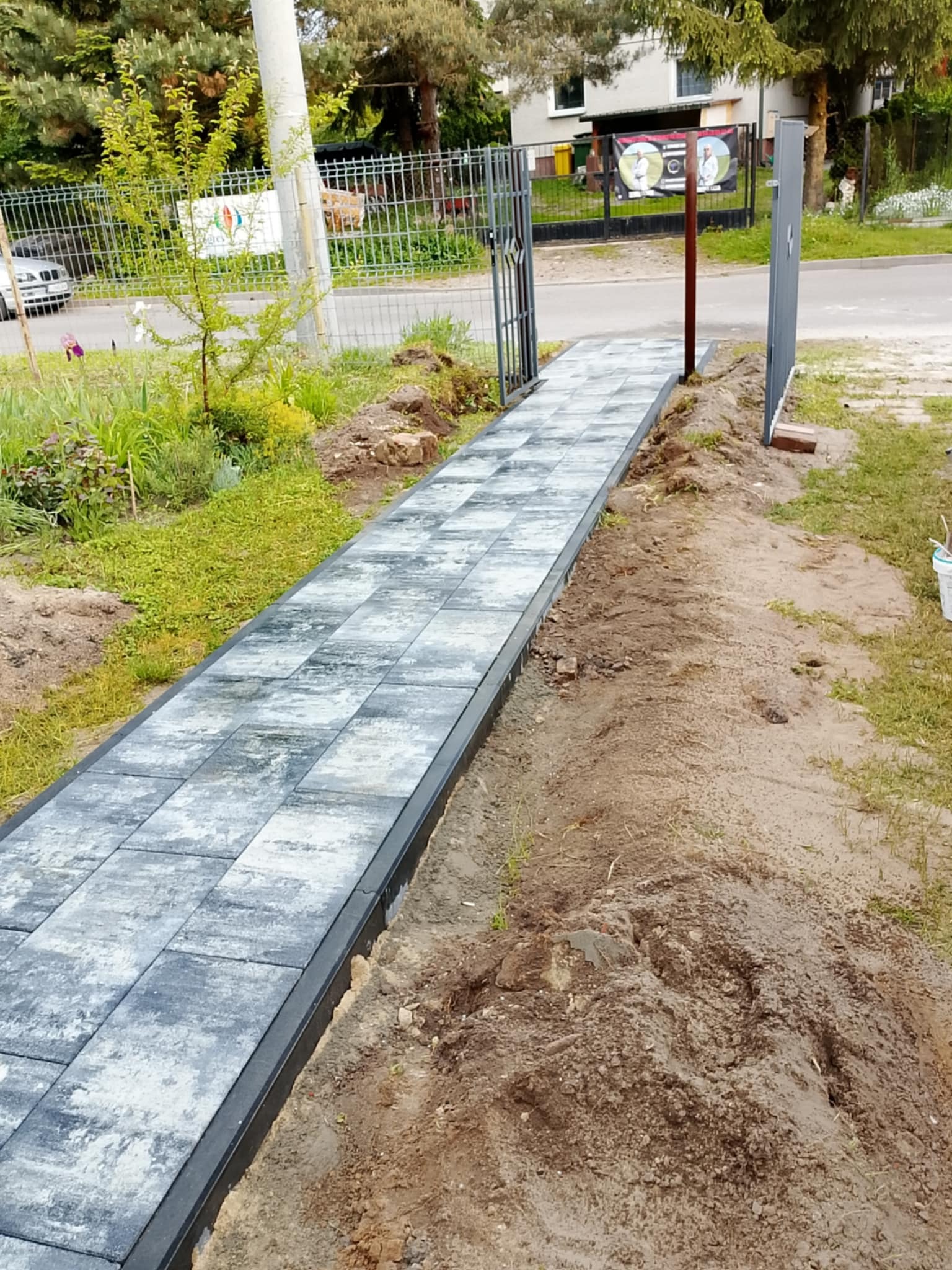 Ścieżka z szarej kostki brukowej prowadząca do bramki ogrodzeniowej. Widoczne świeże obsypanie ziemią. Neutralne oświetlenie, kadr z poziomu gruntu.