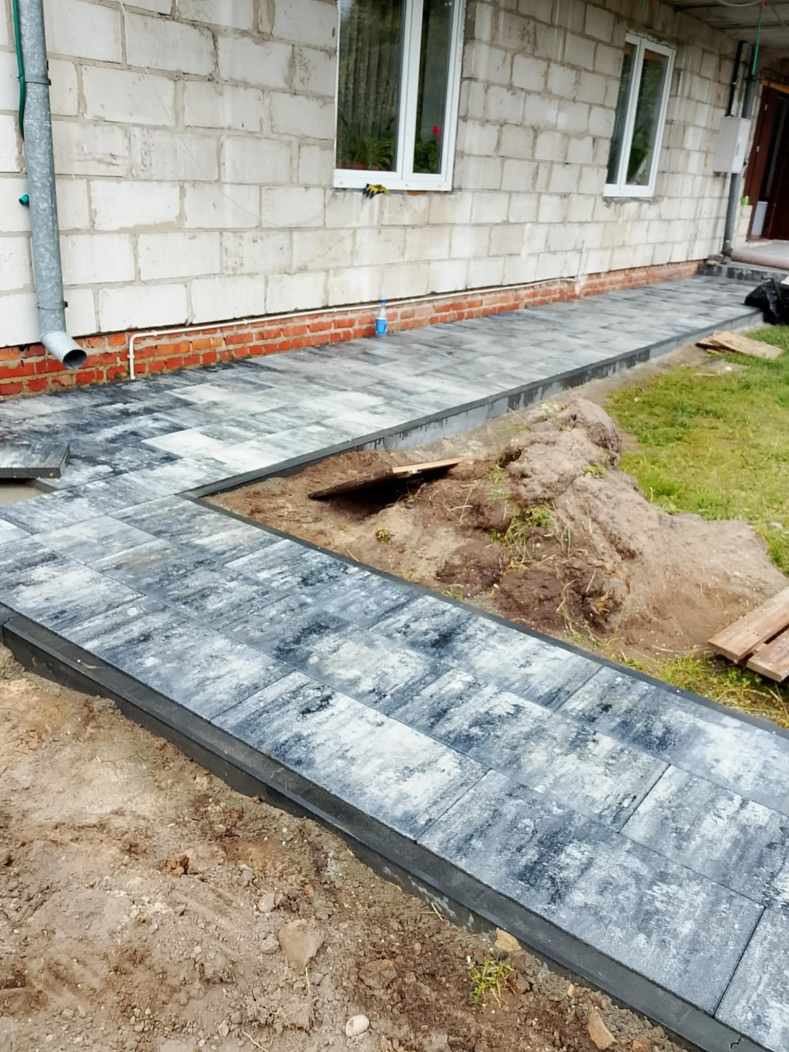 Ścieżka z szarej kostki brukowej ułożona wzdłuż ściany budynku z białego pustaka, z widocznym fragmentem trawnika i przygotowanym podłożem.