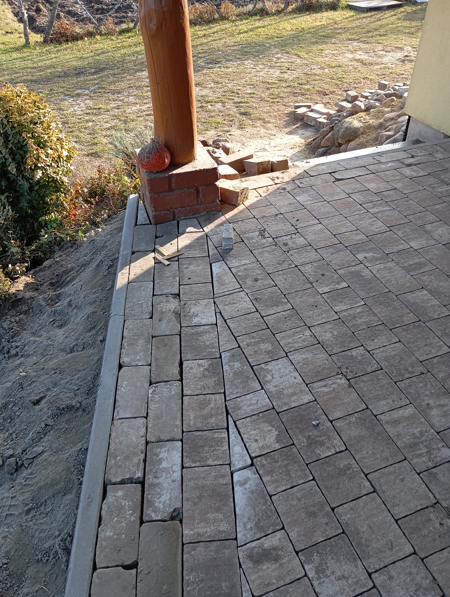 Fragment tarasu z kostki brukowej w trakcie układania, widoczny drewniany słup i przygotowane kostki. Częściowo wykończona powierzchnia w ciepłym świetle.
