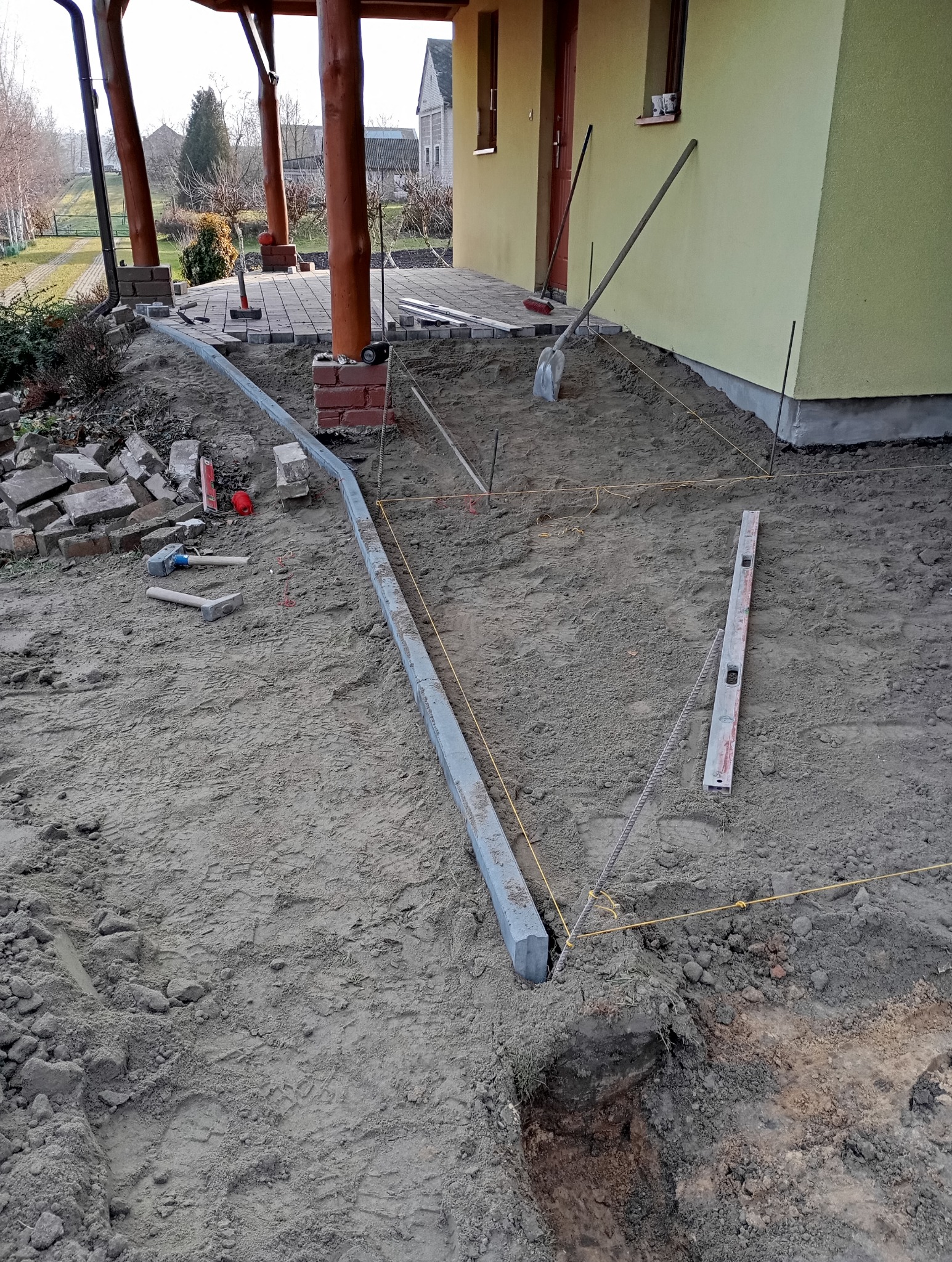 Przygotowanie podłoża pod kostkę brukową: wytyczone granice, poziomica, krawężnik, narzędzia, częściowo ułożona kostka przy wejściu do domu. W tle budynek.