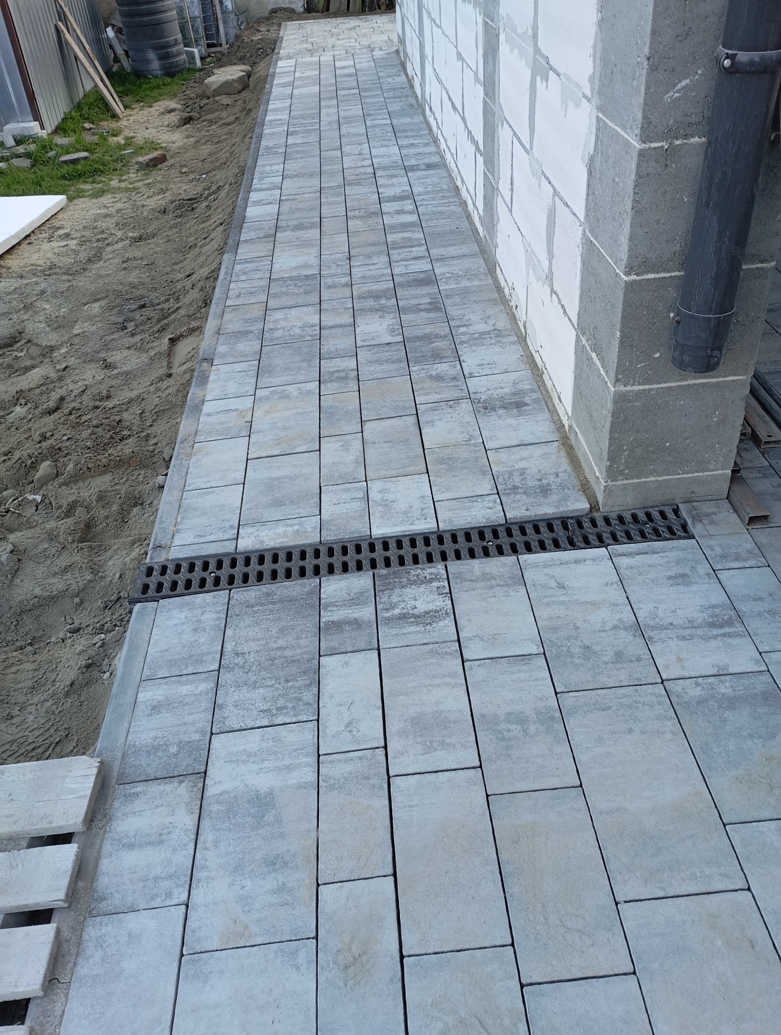 Ułożona szara kostka brukowa na posesji, widoczny odpływ liniowy i fragment ściany budynku z rurą spustową. Prace brukarskie w trakcie realizacji.