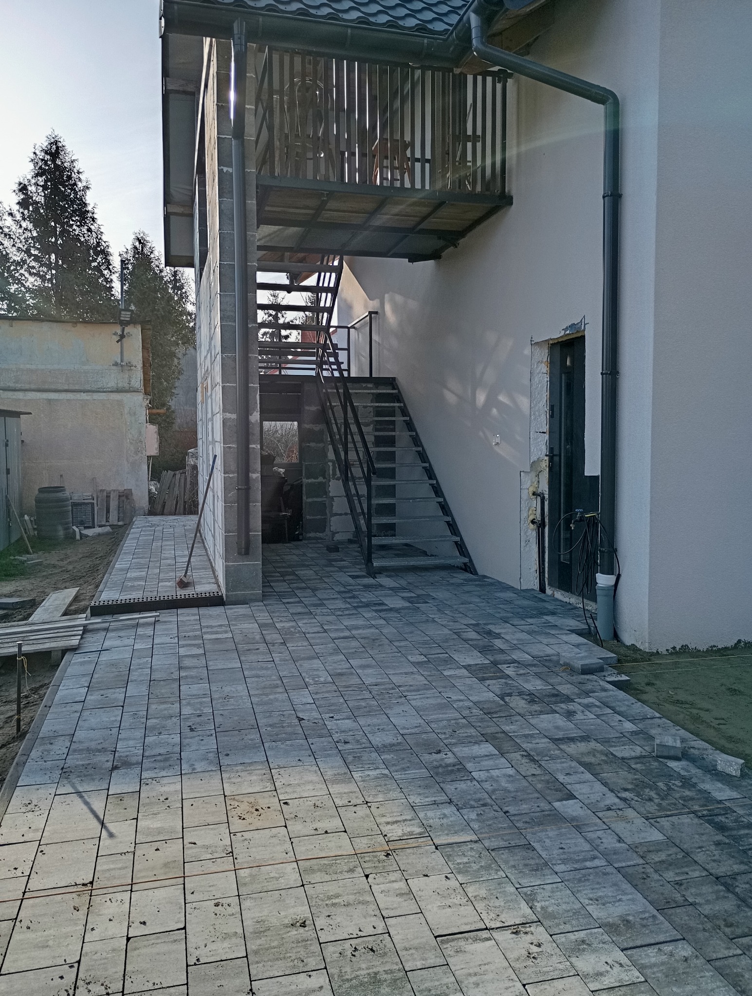 Ułożona szara kostka brukowa na posesji, z widocznymi schodami wejściowymi i fragmentem elewacji budynku. Część kostki jeszcze nieułożona, widoczne narzędzia.