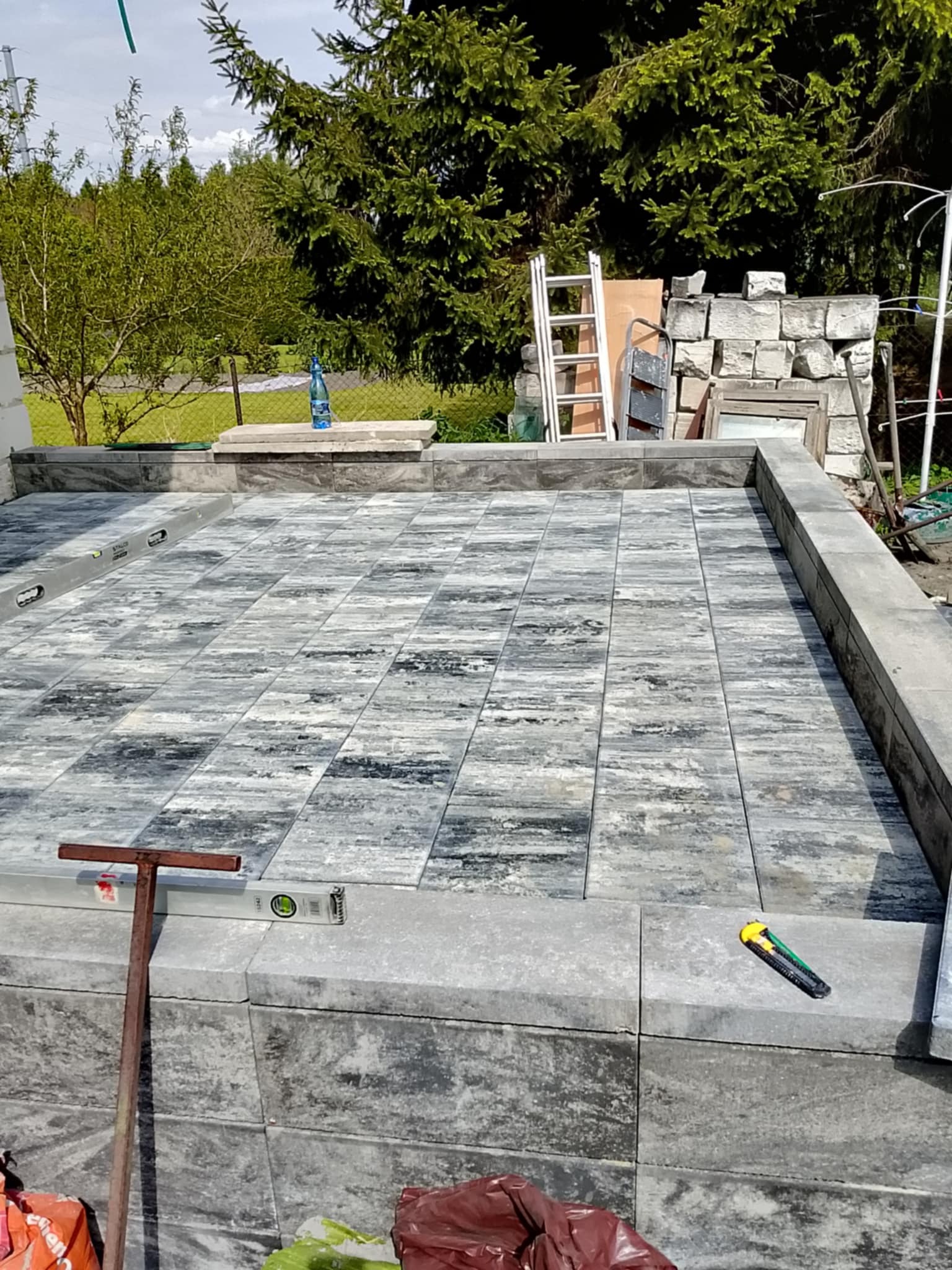 Fragment tarasu z kostki brukowej w odcieniach szarości, otoczony obrzeżami. Widoczne narzędzia: poziomica i nóż. W tle zieleń i elementy konstrukcyjne.