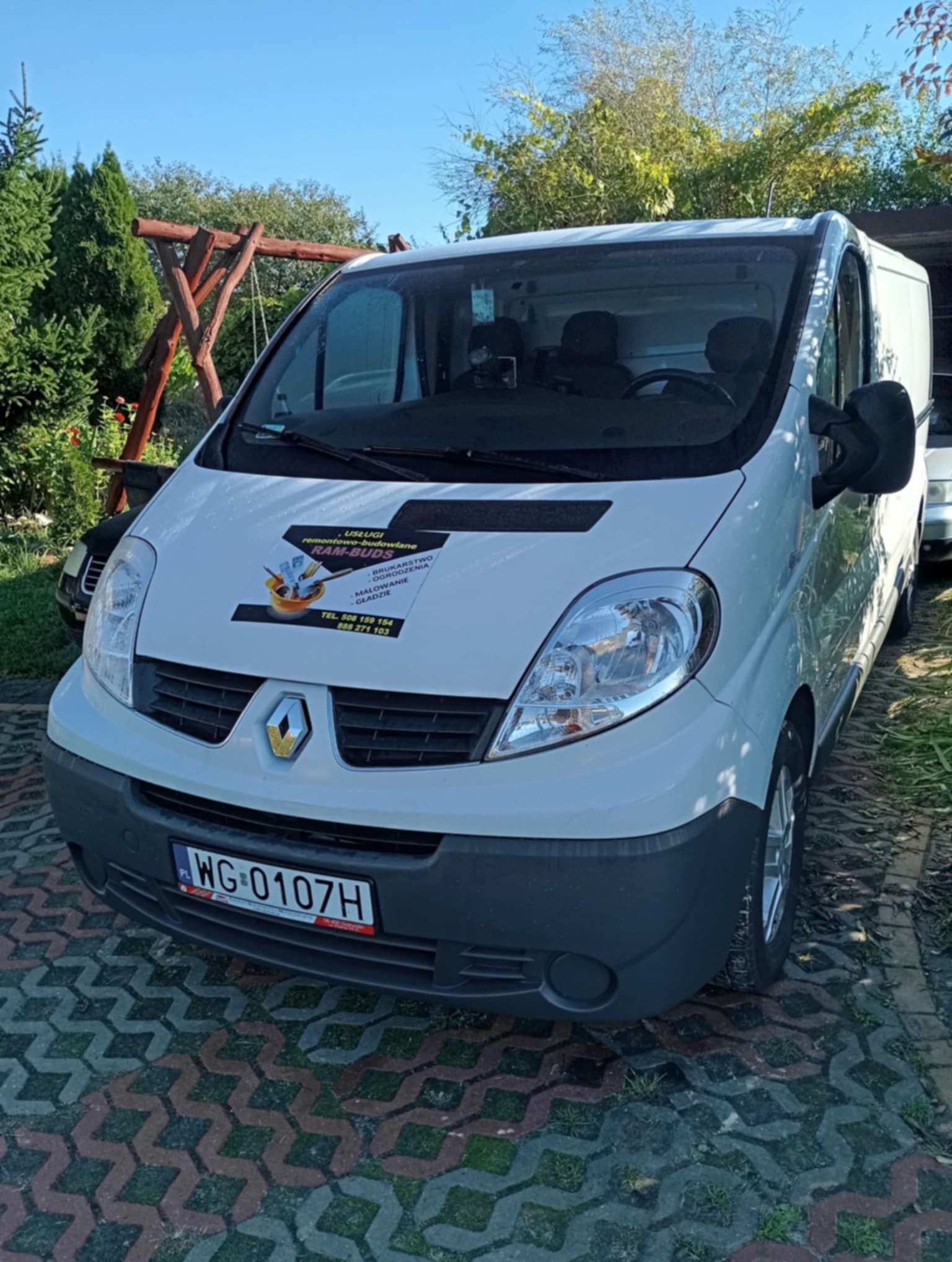 Biały bus firmy RAM-BUDS z naklejką usług remontowo-budowlanych (bruk, ogrodzenia, malowanie) stoi na kostce brukowej. Widoczne numery kontaktowe i logo firmy.