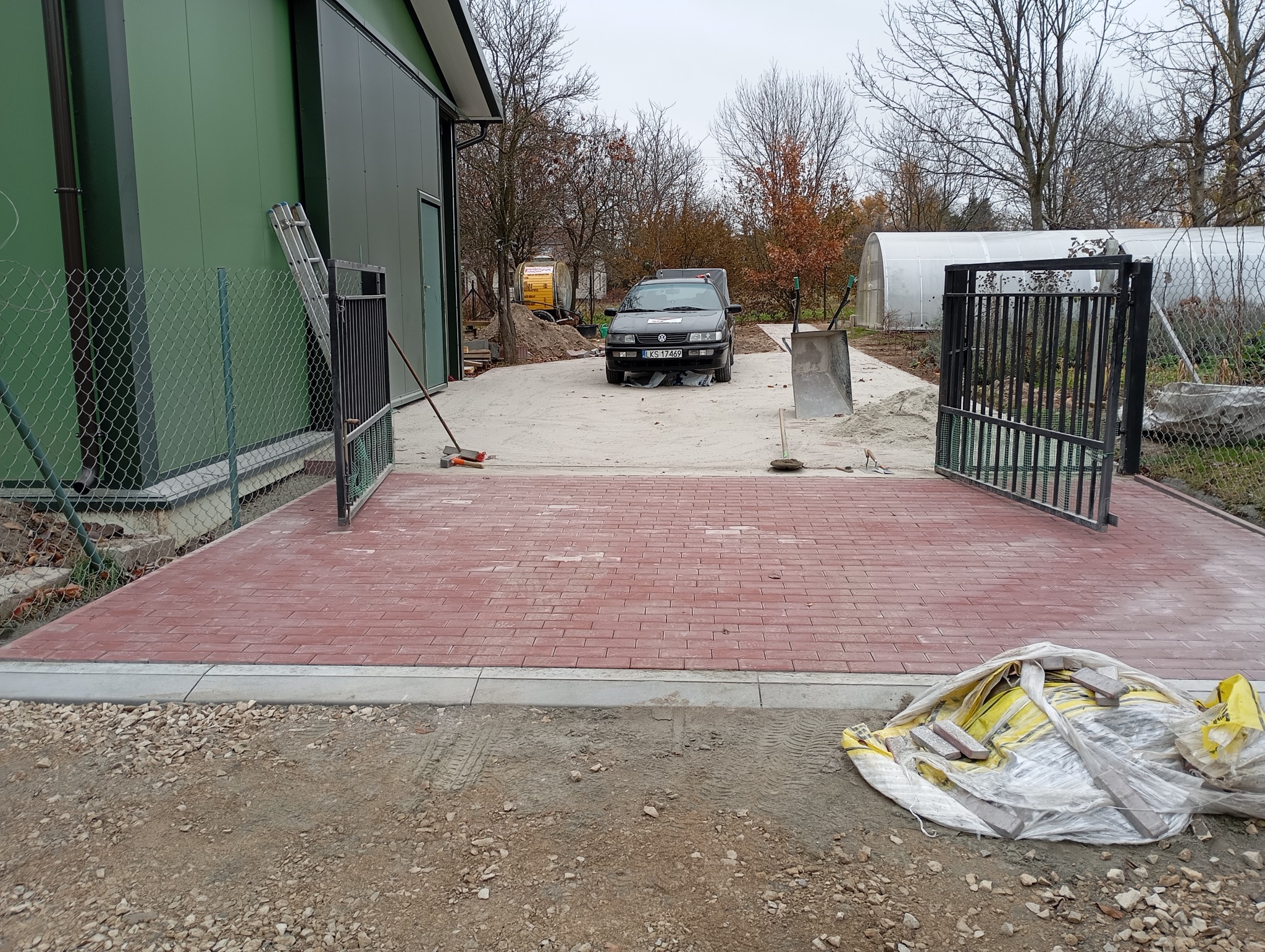 Czerwona kostka brukowa ułożona na podjeździe z otwartą bramą, widoczny samochód i materiały budowlane. Praca w toku, teren przygotowany pod dalsze prace brukarskie.