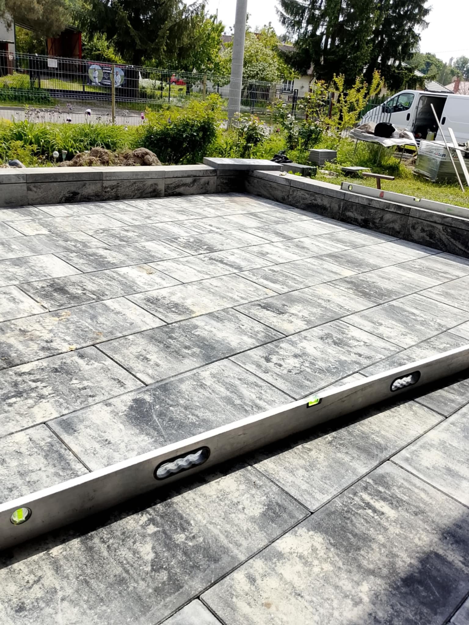 Wykończony taras z kostki brukowej w odcieniach szarości, poziomowany z użyciem poziomicy. W tle zieleń i biały bus.