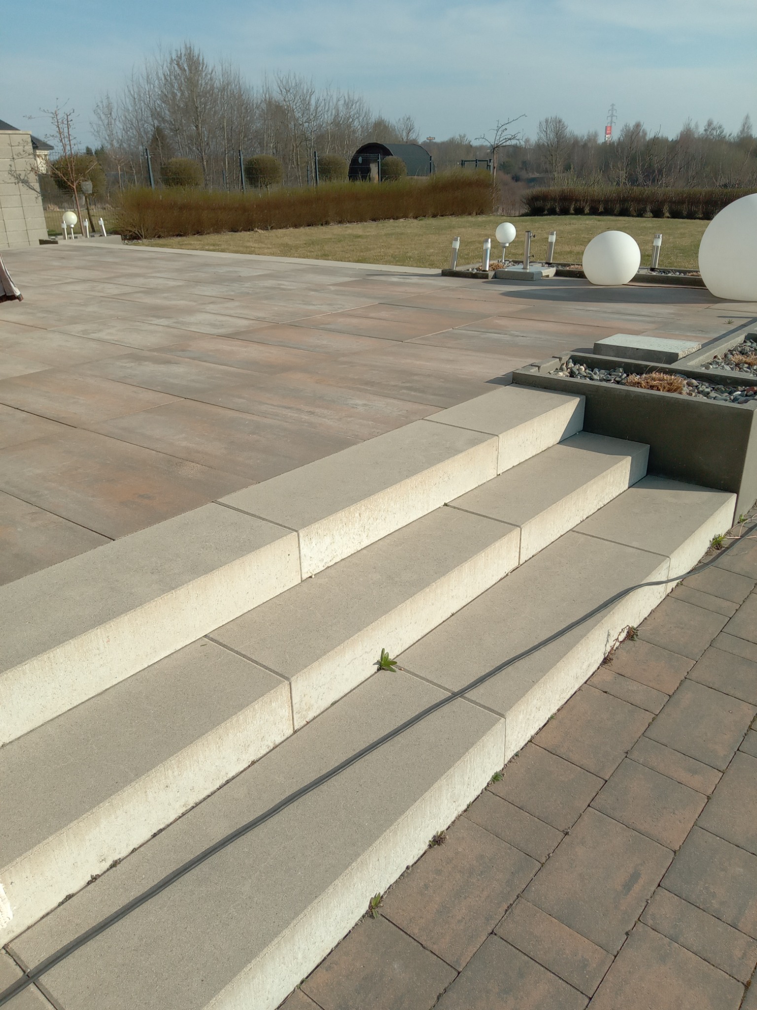 Betonowe schody na tarasie z kostki brukowej w odcieniach brązu. W tle trawnik, krzewy i drzewa. Elementy dekoracyjne w postaci kulistych lamp.