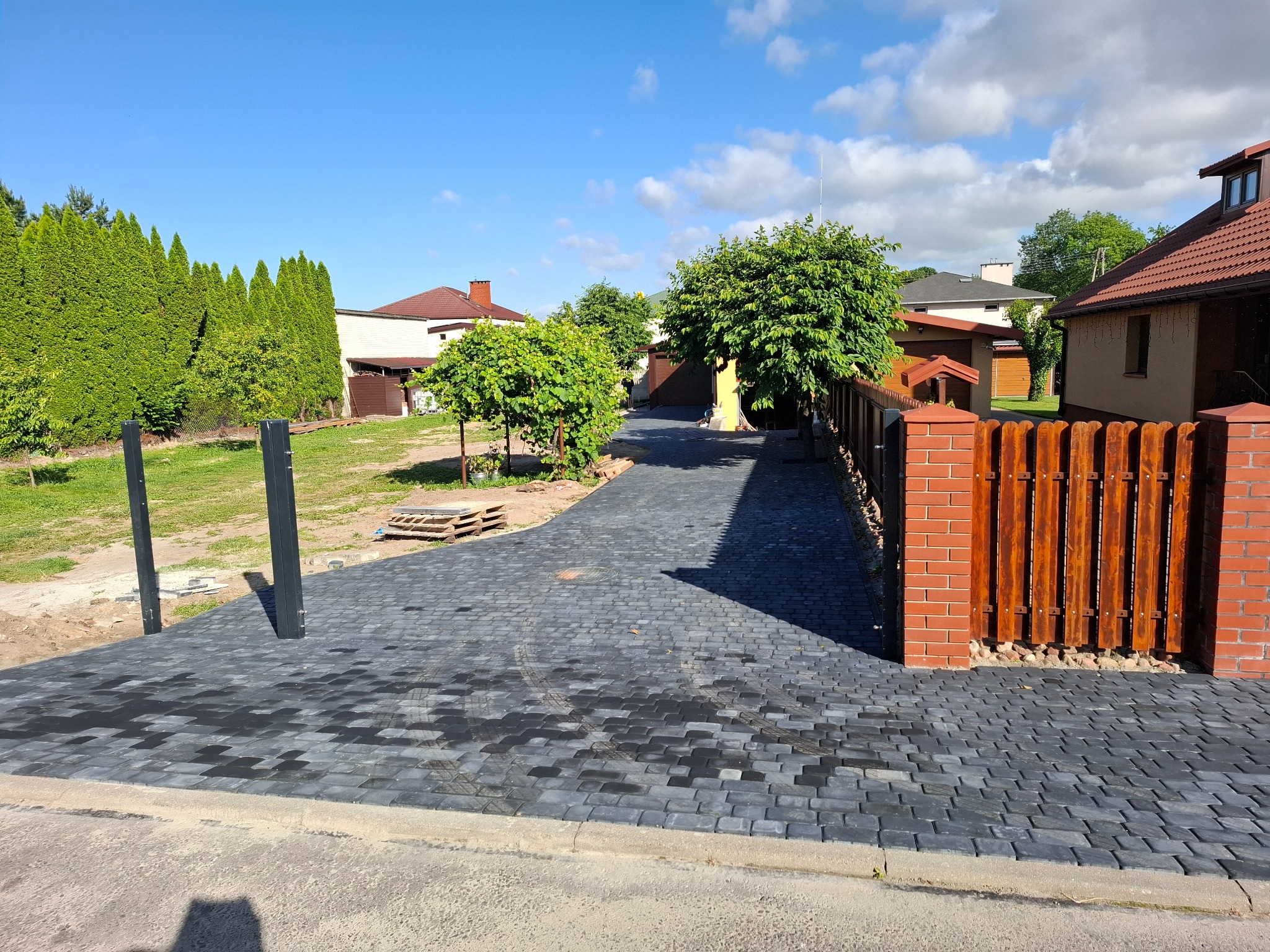 Ułożona, ciemnoszara kostka brukowa prowadząca wzdłuż ogrodzenia z cegły i drewna. Widoczne drzewa, domy w tle i fragment nieba z chmurami.