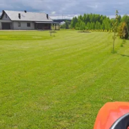 Rozległy trawnik po koszeniu z widocznymi pasami, w tle dom jednorodzinny i drzewa, fragment traktorka do koszenia w kadrze z dołu, pochmurne niebo.