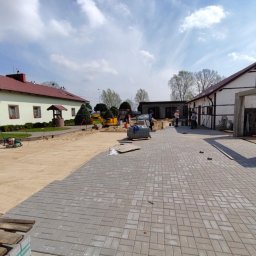 Mirosław Bojar Ogrody Zimowe - Układanie szarej kostki brukowej na posesji. Widoczny mini-koparka, budynki mieszkalne i pracownicy w odblaskowych kamizelkach. Częściowo ułożona nawierzchnia.