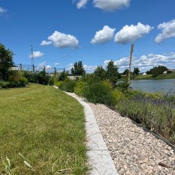 Mirosław Bojar Ogrody Zimowe - Malowniczy ogród z równo przystrzyżonym trawnikiem, kamienną ścieżką i rabatami kwiatowymi nad brzegiem jeziora w słoneczny dzień. Widoczne chmury na niebie.