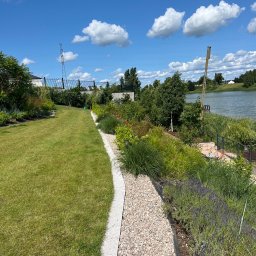 Mirosław Bojar Ogrody Zimowe - Utrzymany trawnik z kamienną ścieżką i rabatami kwiatowymi prowadzącymi do jeziora w słoneczny dzień. Widoczne chmury na niebie i elementy architektury.