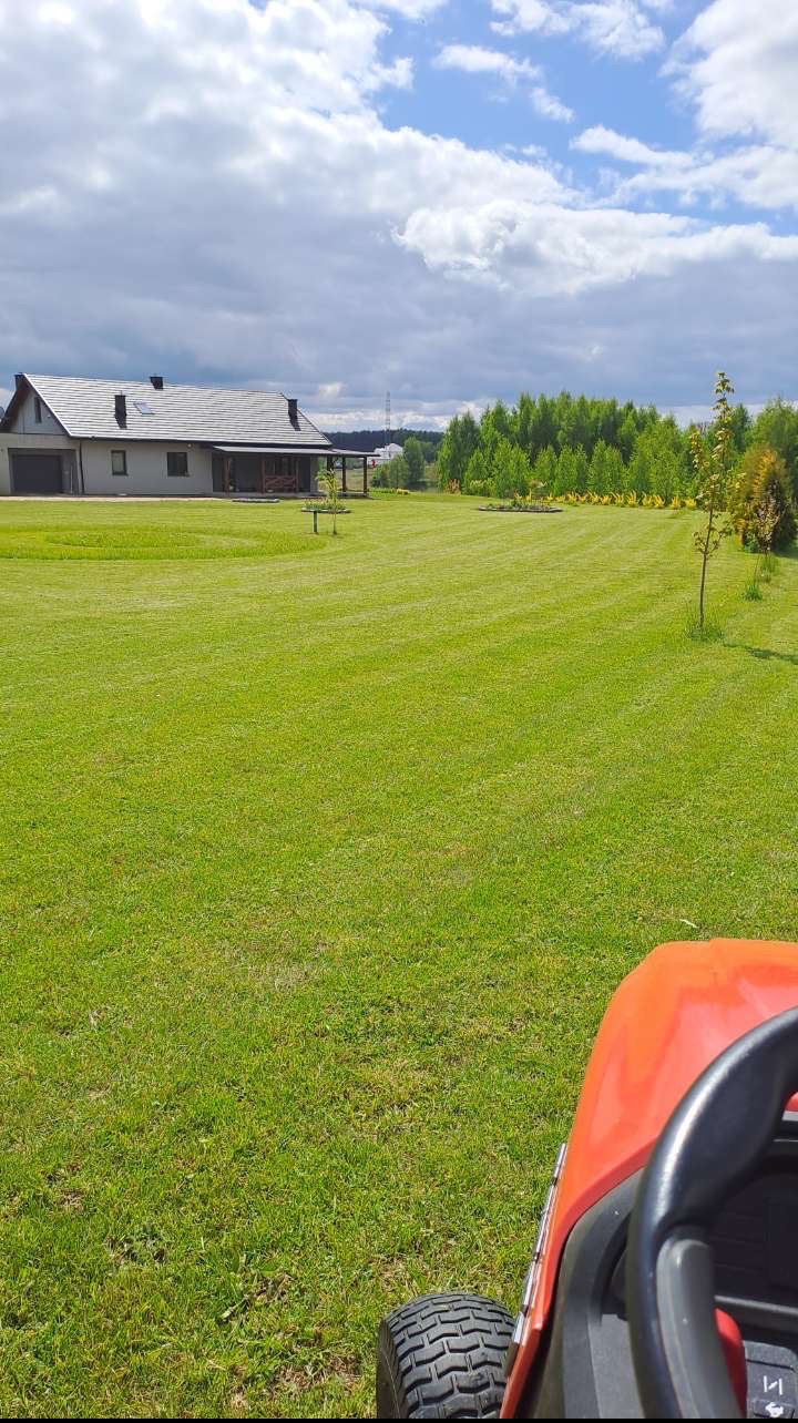 Rozległy trawnik po koszeniu z widocznymi pasami, w tle dom jednorodzinny i drzewa, fragment traktorka do koszenia w kadrze z dołu, pochmurne niebo.