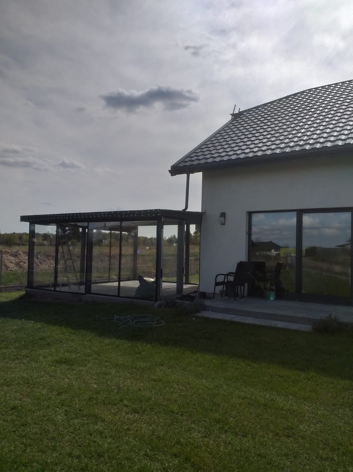 Szklana zabudowa tarasu przylegająca do domu z szarymi oknami i zielonym trawnikiem. Na tarasie drabina i krzesła.