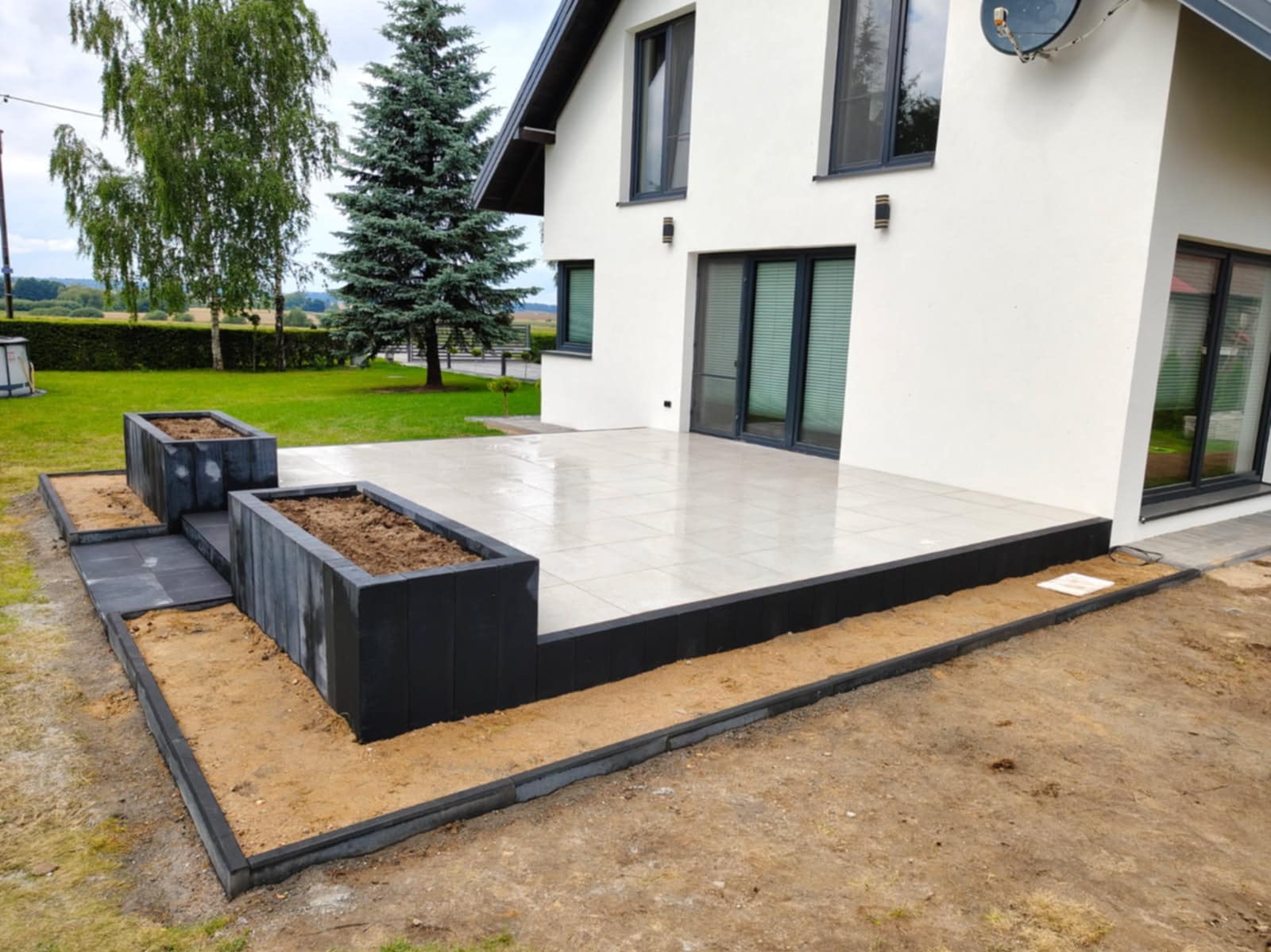 Nowoczesny taras z jasnych płytek i ciemnych obrzeży w Iławie. Donice z ziemią, trawnik i biały dom w tle. Minimalistyczny design.