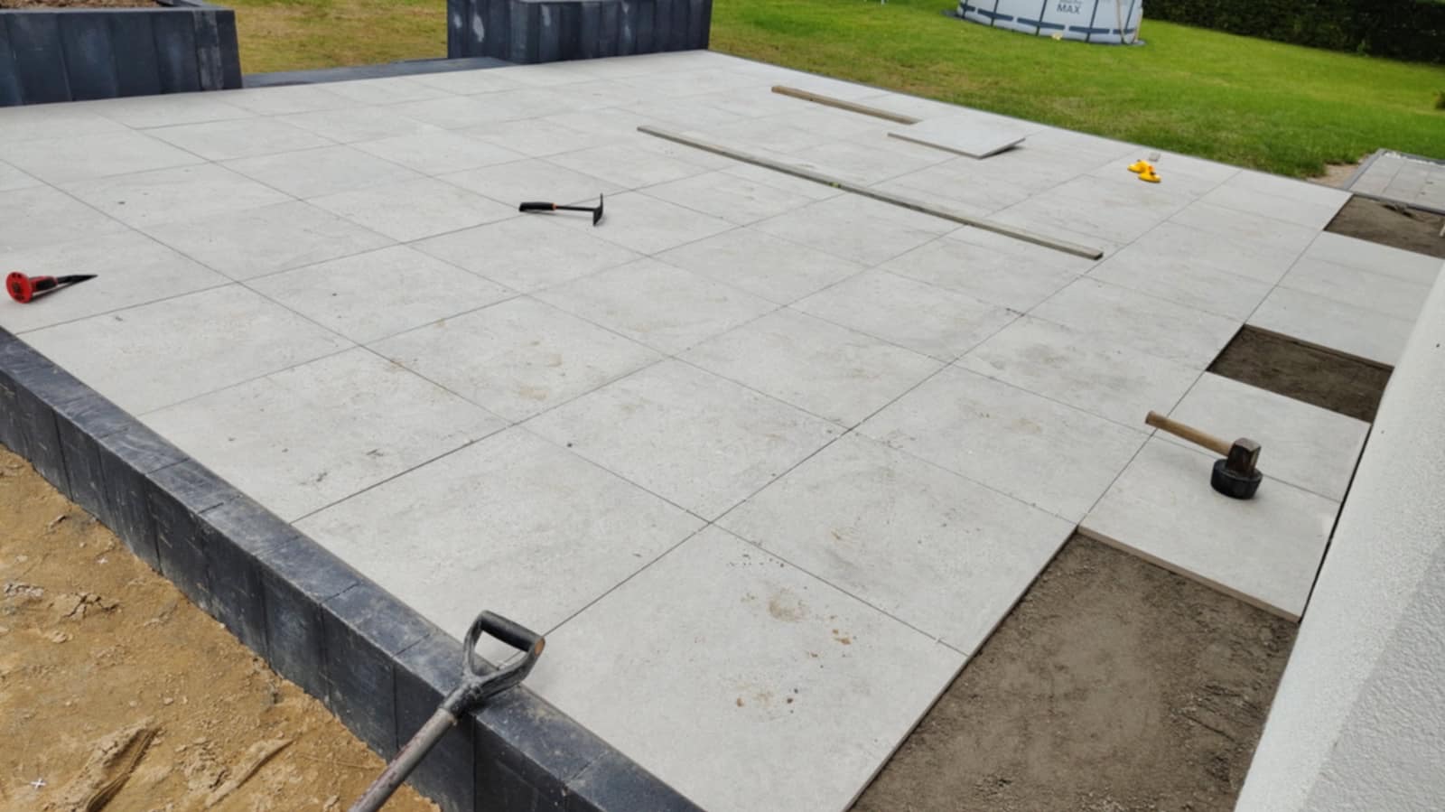 Jasnoszara, kwadratowa nawierzchnia tarasu w trakcie układania. Widoczne narzędzia: młotek, poziomica, łopata. Część płyt brakuje, ukazując podłoże.