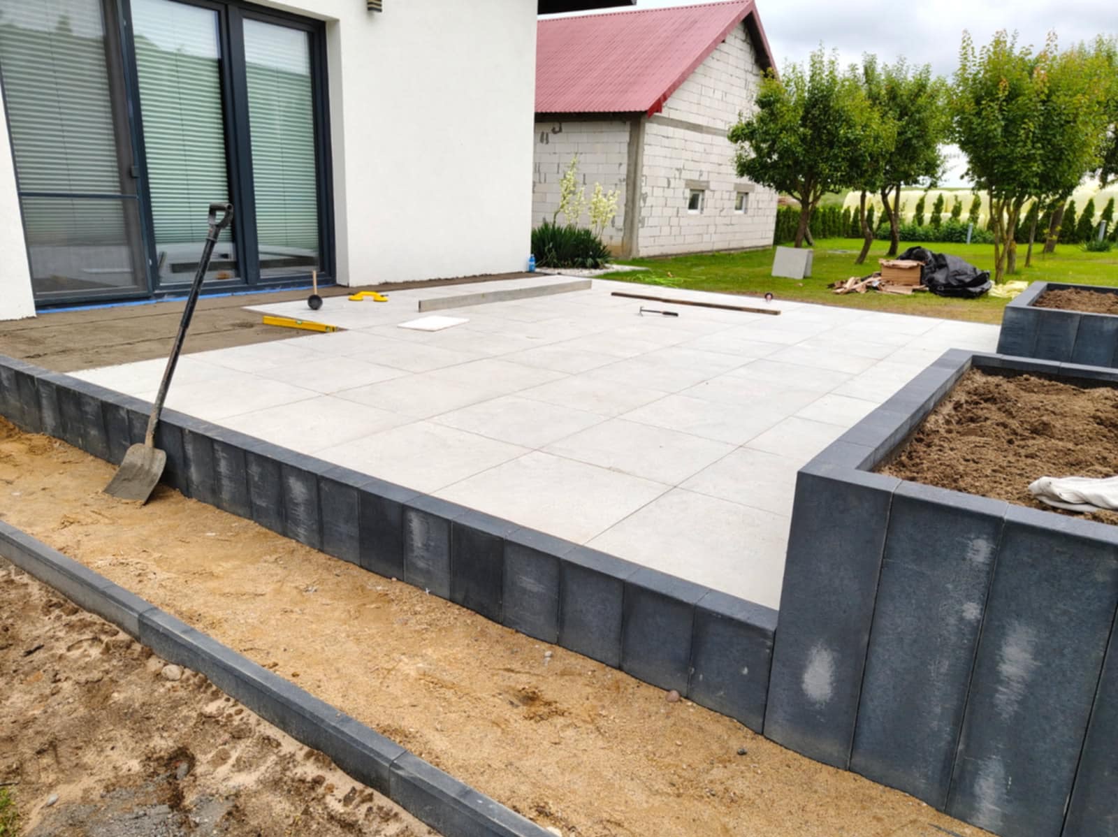 Jasnoszara, kwadratowa kostka brukowa ułożona na tarasie, obrzeża z ciemnoszarych elementów, widoczna łopata i poziomica, w tle dom z czerwonym dachem i zieleń.