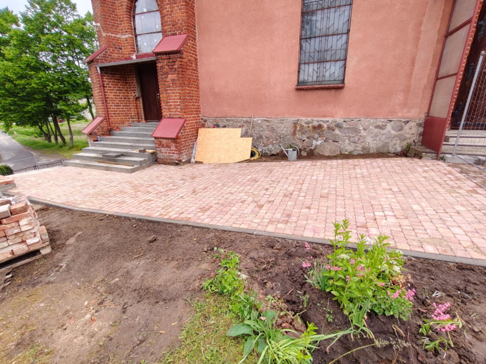 Ułożona kostka brukowa w odcieniach czerwieni przed budynkiem z cegły. Widoczne przygotowane rabaty kwiatowe i narzędzia. Częściowo widoczny stos cegieł.