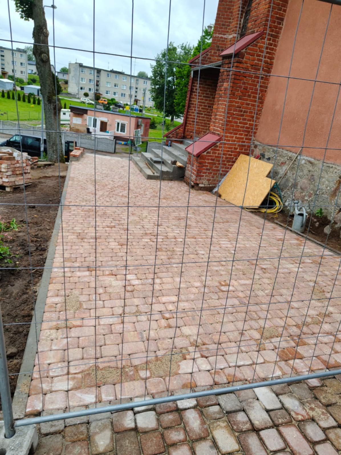 Ułożona, czerwona kostka brukowa tworząca ścieżkę prowadzącą do schodów budynku. Widoczne narzędzia i materiały budowlane. Całość ogrodzona siatką.