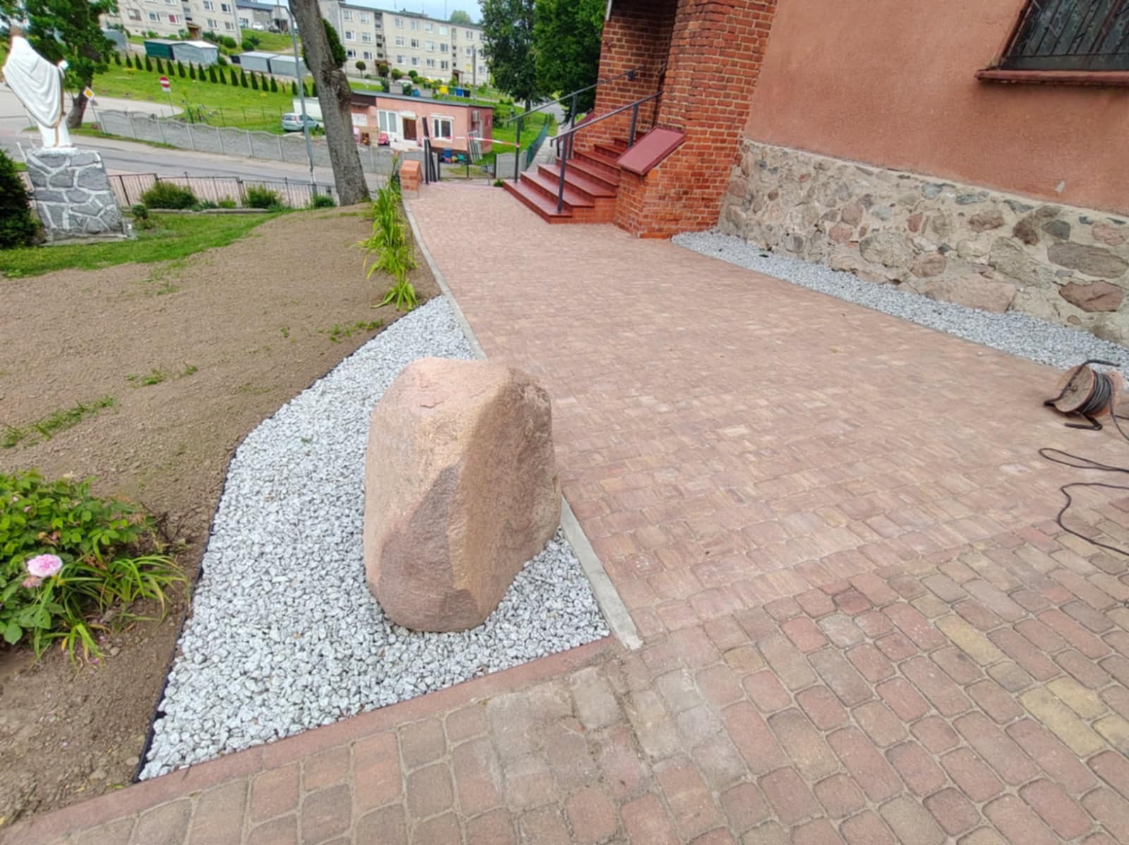 Ułożona kostka brukowa w odcieniach czerwieni tworząca ścieżkę, obok rabata z kamieniami i dużym głazem. W tle budynek z cegły i schody.