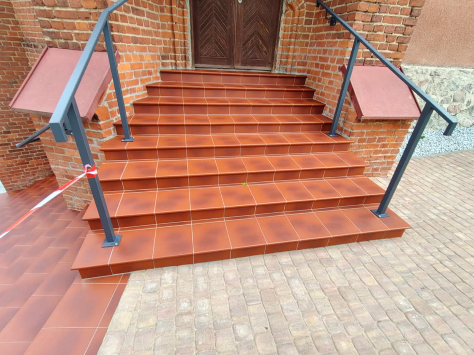 Kaskadowe schody z terakotowej płytki, wykończone szarą metalową balustradą, prowadzące do drewnianych drzwi w ceglanej fasadzie. Widoczna kostka brukowa.