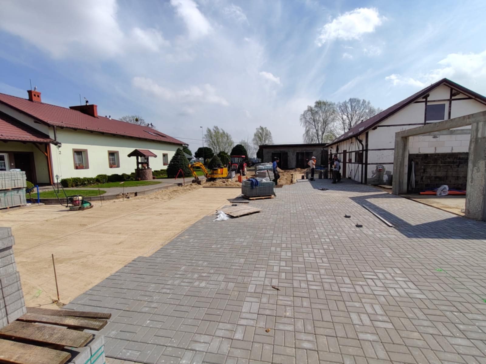 Układanie szarej kostki brukowej na posesji. Widoczny mini-koparka, budynki mieszkalne i pracownicy w odblaskowych kamizelkach. Częściowo ułożona nawierzchnia.