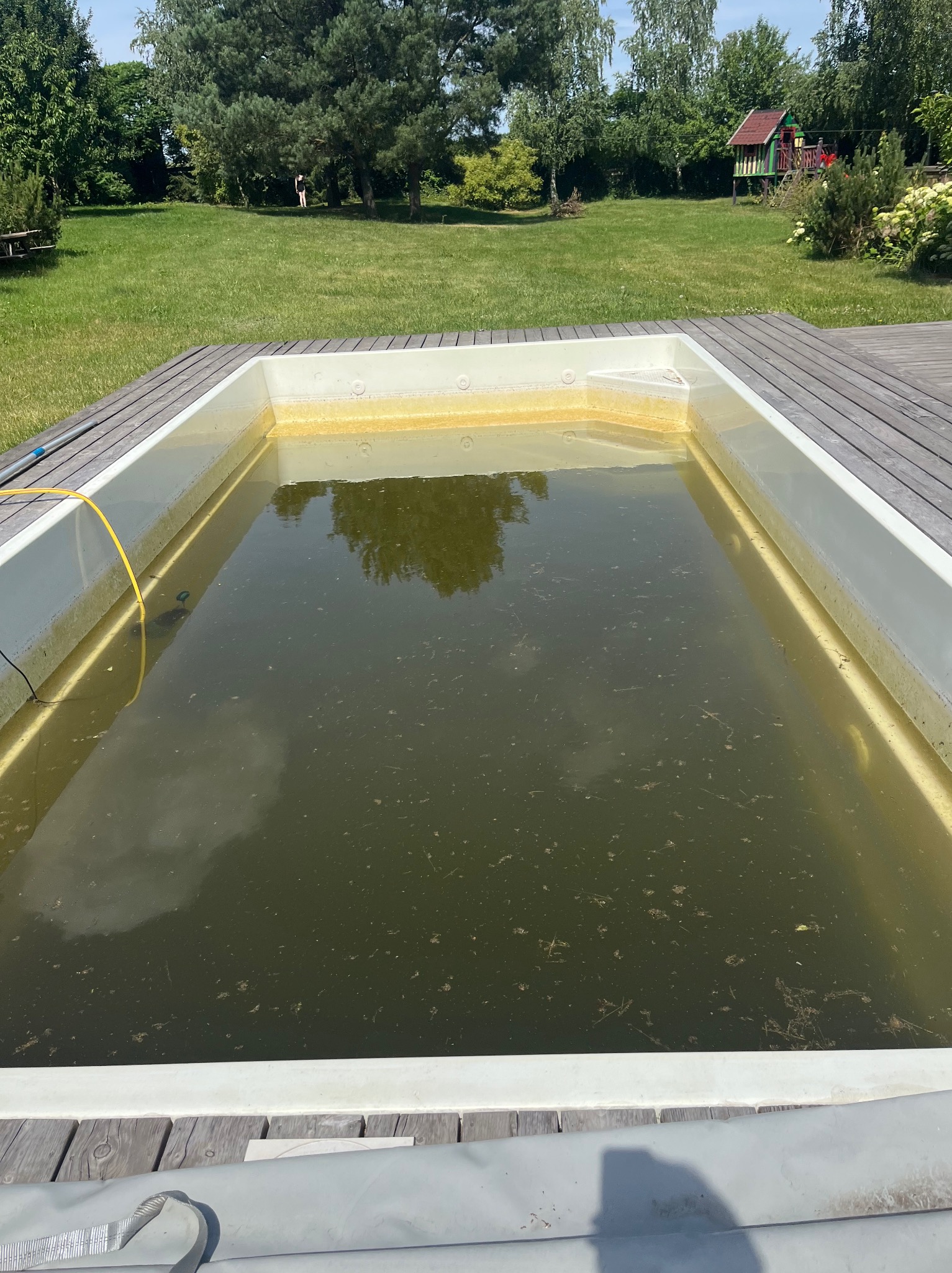 Zanieczyszczony basen ogrodowy z zielonkawą wodą, otoczony drewnianym tarasem. W tle trawnik, drzewa i domek dla dzieci. Widoczne osady na ścianach basenu.