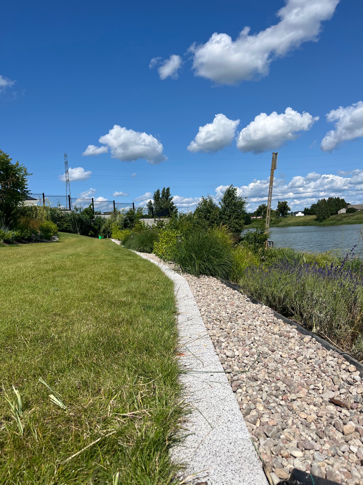 Malowniczy ogród z równo przystrzyżonym trawnikiem, kamienną ścieżką i rabatami kwiatowymi nad brzegiem jeziora w słoneczny dzień. Widoczne chmury na niebie.
