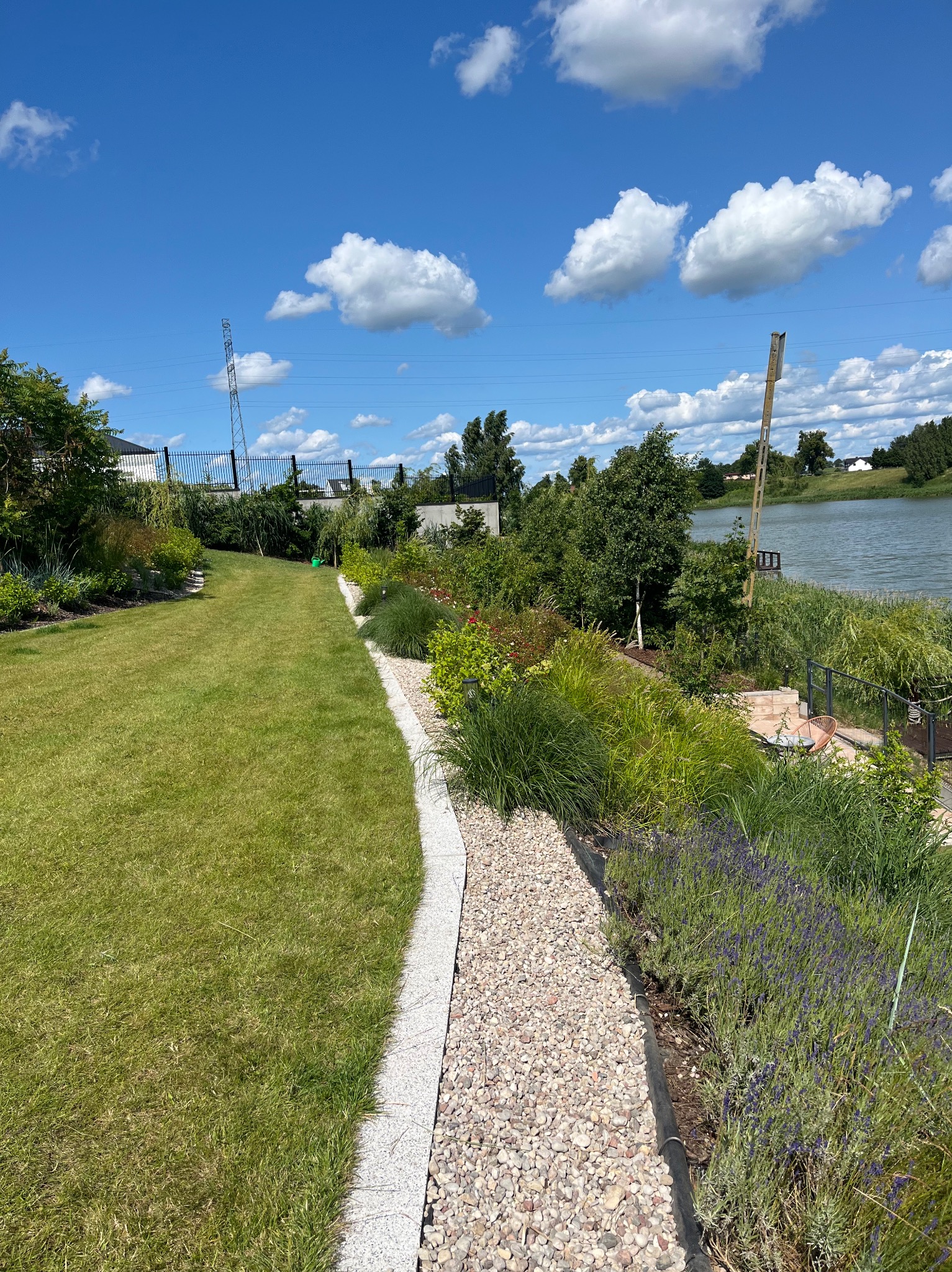 Utrzymany trawnik z kamienną ścieżką i rabatami kwiatowymi prowadzącymi do jeziora w słoneczny dzień. Widoczne chmury na niebie i elementy architektury.