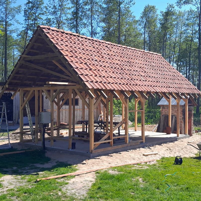 Wznoszona altana z drewnianą konstrukcją i częściowo ułożonym czerwonym dachem z dachówki, na tle zieleni i drzew. Widoczne narzędzia i elementy budowlane.