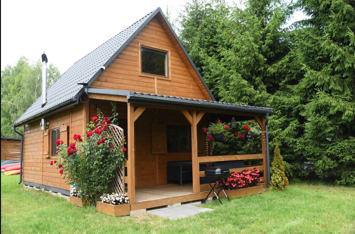Drewniany domek letniskowy z zadaszonym tarasem, ozdobiony kwitnącymi różami i kwiatami w donicach. Szara dachówka, komin i zielone otoczenie drzew.