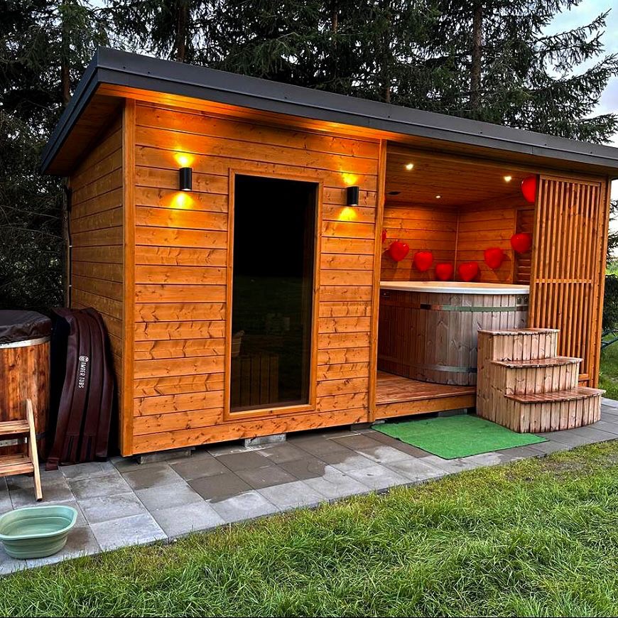 Drewniana altana z jacuzzi, oświetlona lampami ściennymi i dekoracjami w kształcie serc. Schodki prowadzą do wnętrza, a obok widoczna wanna i zielony trawnik.