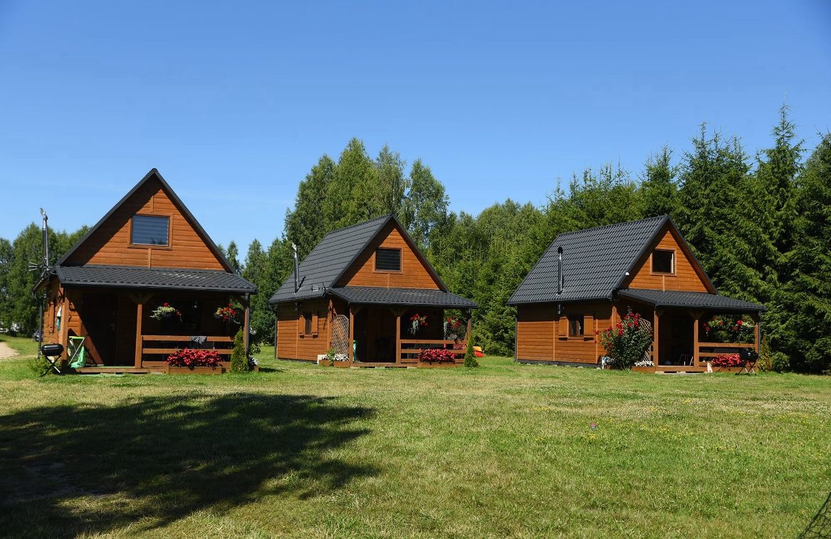 Trzy drewniane domki letniskowe z ciemnymi dachami na zielonej trawie, otoczone drzewami. Domki mają małe werandy z kwiatami, widoczne kominy i okna na poddaszu.