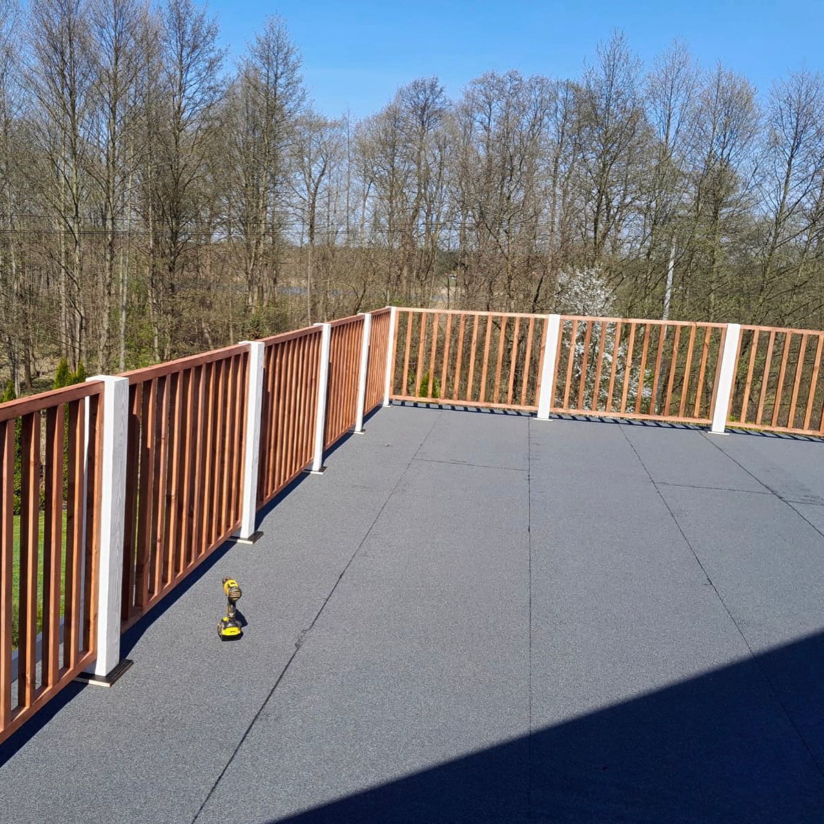 Nowoczesny taras z drewnianą balustradą i szarą, gumową nawierzchnią. W tle drzewa i błękitne niebo. Wiertarka akumulatorowa leży na podłodze tarasu.