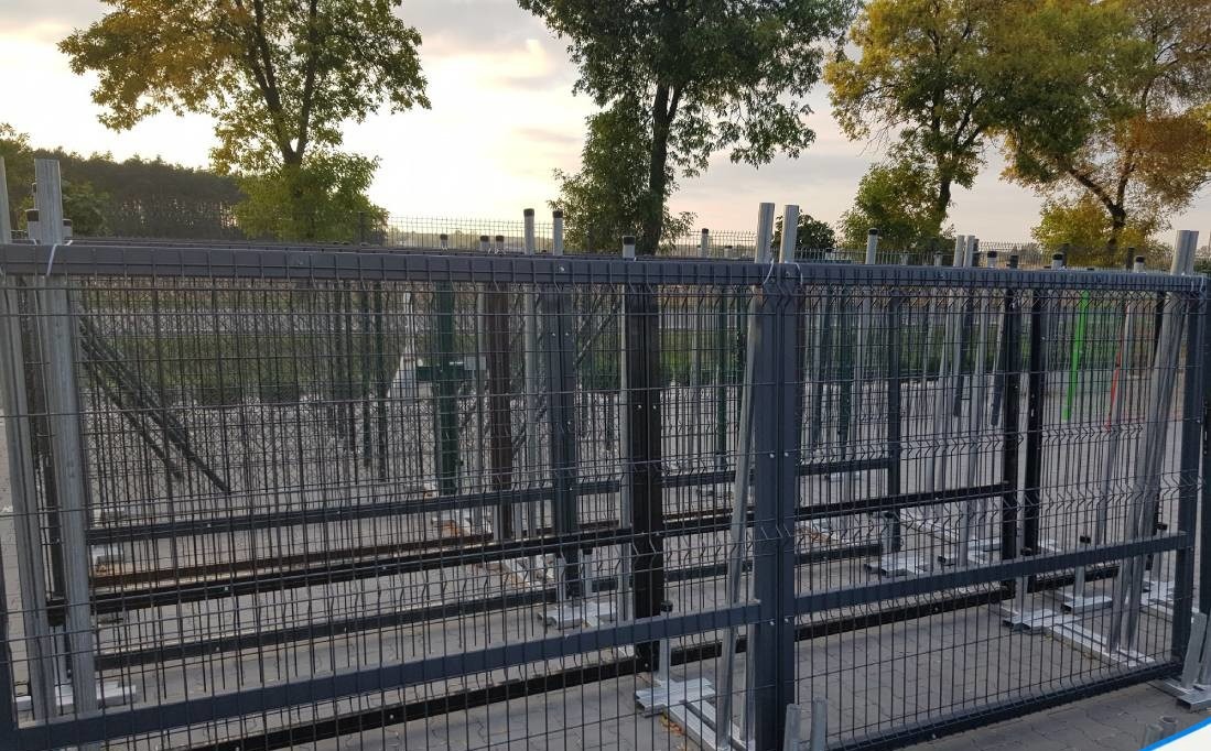 Szare panele ogrodzeniowe z metalowymi słupkami montowane na betonowej podstawie, widoczne drzewa w tle.