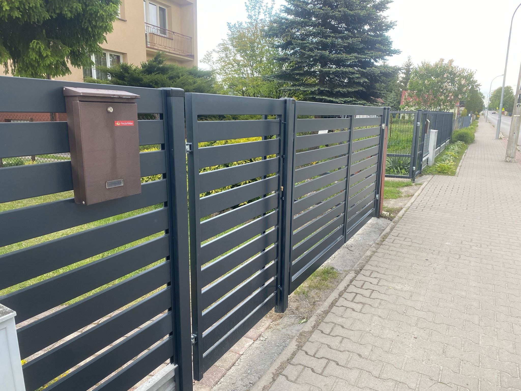 Nowoczesne, poziome przęsła ogrodzeniowe w antracytowym kolorze, z brązową skrzynką pocztową. Widok z chodnika na tle zieleni i budynku.