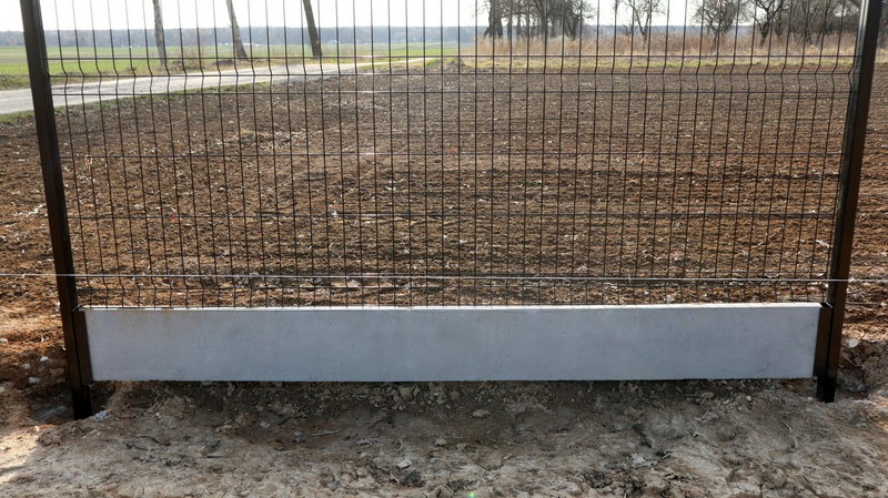 Fragment montażu panelowego ogrodzenia z podmurówką betonową na tle zaoranej ziemi i drzew. Widoczne słupki i siatka panelowa.