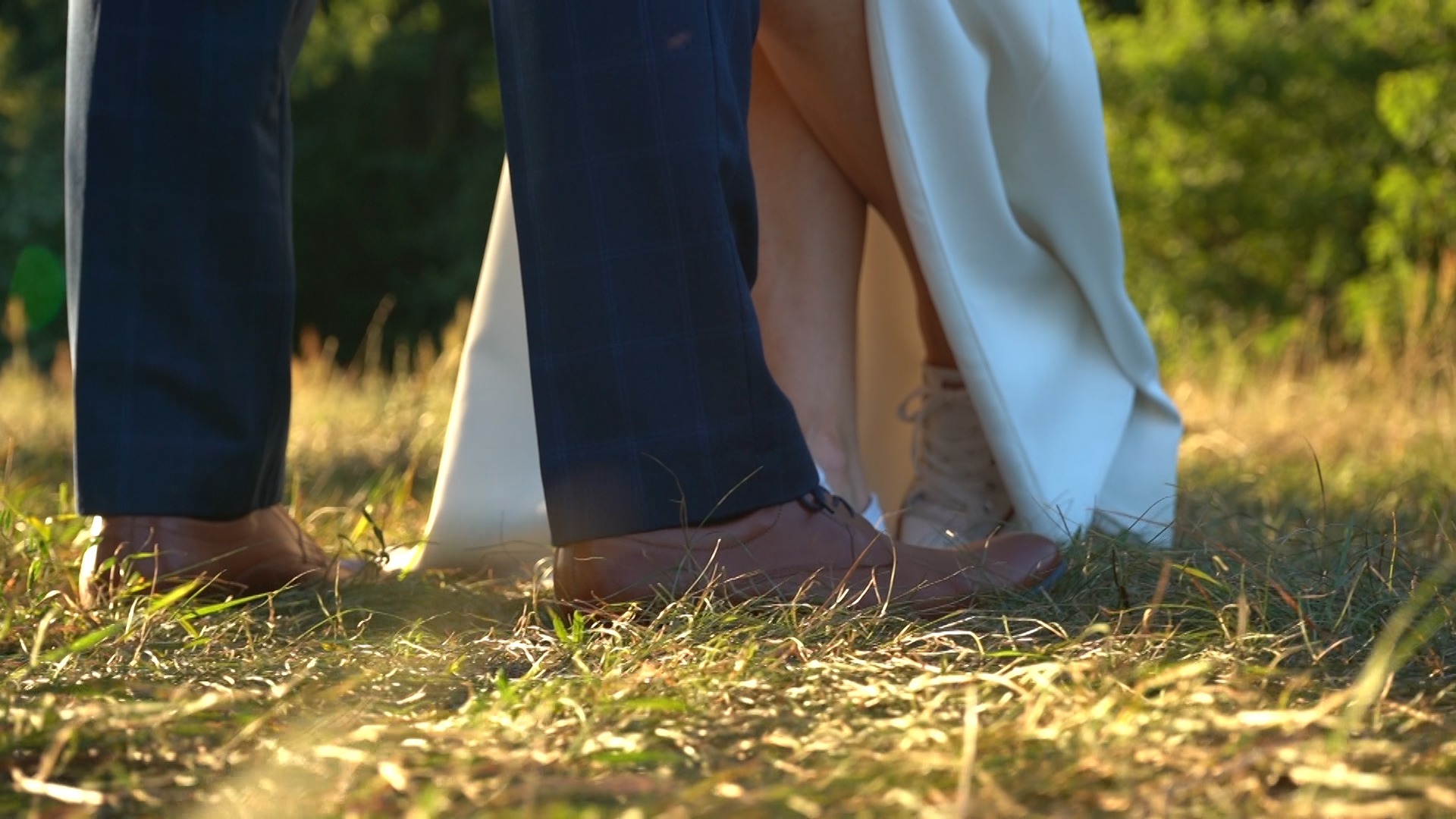 Fragment pary młodej w plenerze: nogi mężczyzny w brązowych butach i kobiety w białej sukni i beżowych butach sportowych na tle trawy w promieniach zachodzącego słońca.